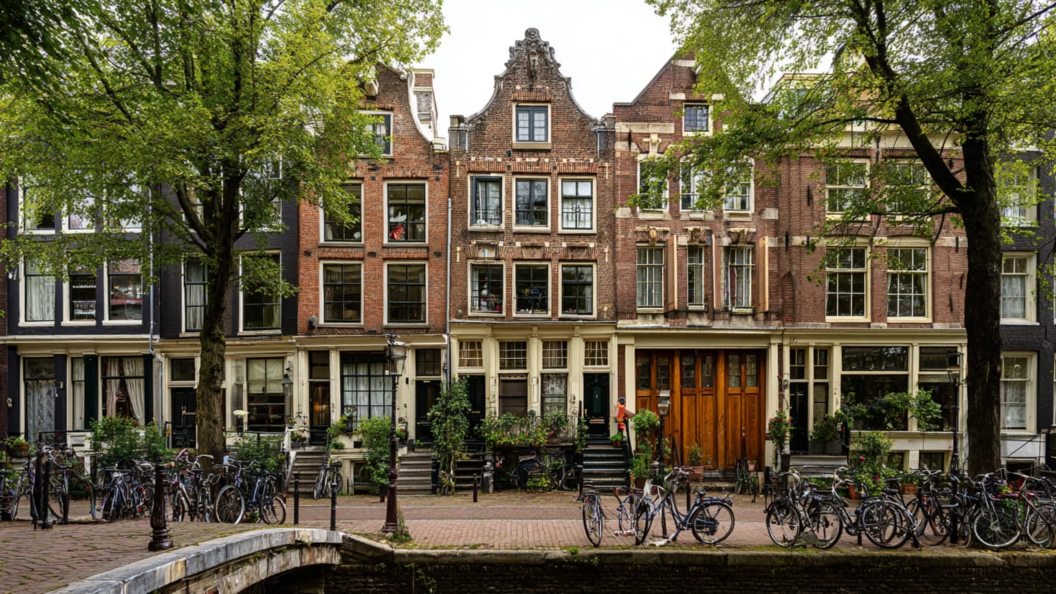 Evening light on an Amsterdam canal with bicycles, brick row houses, trees, and people walking along the water
