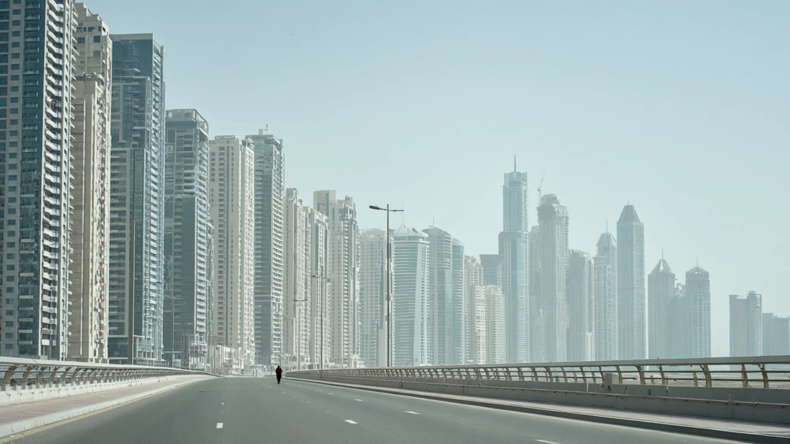 A Dubai boulevard at twilight with towers, wide roads, a metro line, and small groups of pedestrians moving through the heat